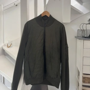 J Lindeberg cardigan  - Snygg olivgrön jacka från J.Lindeberg med quiltad front och stickade ärmar. Jackan har hög krage, hel dragkedja och två fickor med dragkedja. Perfekt för dig som gillar stilren design med sportig touch.