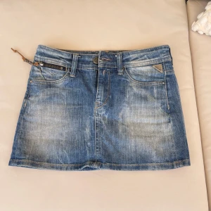 Blå jeanskjol från Replay - Snygg blå jeanskjol från Replay med klassisk femficksdesign, slitna detaljer och coolt broderat Replay-märke med ros på baksidan. Kjolen har dragkedja, knapp och rutiga innerfickor som ger extra edge. Perfekt till sneakers eller boots.
