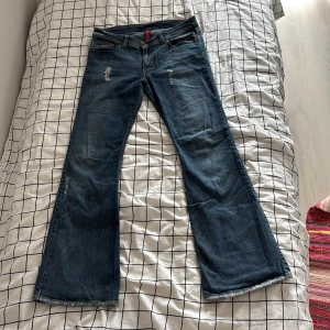 Blå bootcut jeans med broderade fickor - Snygga blå bootcut jeans från H&M med slitna detaljer framtill och coolt broderi på bakfickorna. Klassisk femficksmodell i jeansmaterial med låg midja och lätt tvättad look. Byxorna har slitningar men inget som syns när man har på sig dem. Midjemåttet är 88 cm runt och innerbenslängden är 78 cm. Byxorna är vintage från hm