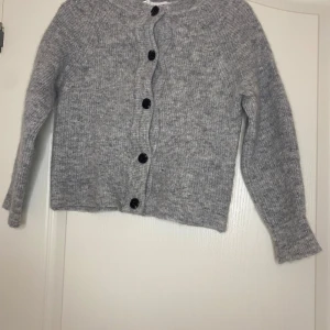 Grå stickad kofta från Selected Femme - Mysig grå stickad kofta från Selected Femme med rund halsringning och svarta knappar framtill. Koftan har långärmad passform och är tillverkad i ett fluffigt, mjukt material som passar perfekt för kyliga dagar. Enkel och stilren design som är lätt att matcha.