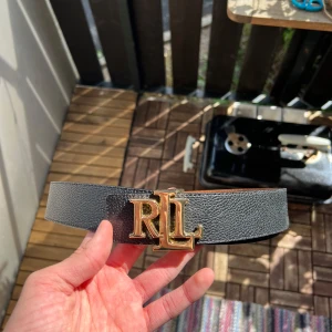Svart skärp från Ralph Lauren (Guld) - Stilrent svart bälte från Ralph Lauren med bred rem i präglat skinn och en stor, guldig RL-logga som spänne. Perfekt accessoar för att lyfta vilken outfit som helst med en lyxig touch.