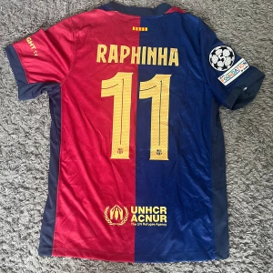  Barcelona Raphinha #11 Nike fotbollströja - FC Barcelona matchtröja med Raphinha och nummer 11 på ryggen. Tröjan är kortärmad, i polyester, med klassisk röd och blå halvdelad design, guldiga detaljer, klubbmärke, Nike-logga, Spotify och UNHCR-tryck samt Champions League-märke på ärmen.