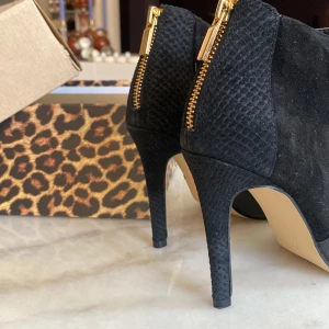 Svarta klackar med guldkedja  - Svarta klackar  från River Island med spetsig tå, hög klack och guldfärgade dragkedjor bak. Skorna har en mix av slätt skinn och mocka med strukturdetaljer på hälen.  Kartongen ingår med 