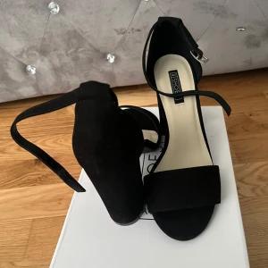 Svarta klackade sandaler från NLY Shoes - Stilrena svarta sandaler från NLY Shoes med bred klack och öppen tå. Skorna har en smal rem runt vristen med silverfärgat spänne och är tillverkade i mockaliknande material. Perfekta för att lyfta din outfit med en clean och modern vibe. HR bara testa de när jag fick de insåg att de var för höga för mig och för stora