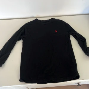 Svart långärmad tröja Polo Ralph Lauren - Titel: Svart långärmad tröja från Ralph Lauren 🖤  Beskrivning: Supersnygg långärmad tröja från Ralph Lauren i svart med klassisk röd logga på bröstet. Passar perfekt till både chill och mer uppklädda outfits! Skön passform och tidlös stil. Det är två pyttesmå hål som man knappt märker av.  ✔️ Äkta Ralph Lauren ✔️ Röd broderad logga. Nypris ca 1000 kr – säljer pga garderobsrensning.  💬 Fråga gärna om fler bilder!