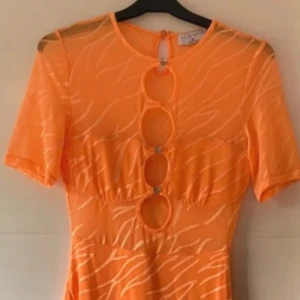 Orange kortklänning med cutouts - Snygg orange kortklänning med zebramönster och tre runda cutouts framtill, dekorerade med små glittriga stenar. Klänningen har korta ärmar och är gjord i ett lätt, syntetiskt material som känns luftigt. Perfekt för dig som vill sticka ut med coola detaljer och färg. Det saknas en knapp, men man kan ta bort dom andra och behålla ena uppe. Använd en gång.