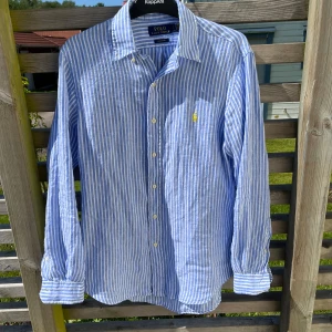 Blårandig linneskjorta Polo Ralph Lauren - Snygg blårandig skjorta från Polo Ralph Lauren i luftig linne. Klassisk krage, långa ärmar och knäppning framtill. Custom fit-modell med ikonisk gul broderad logga på bröstet. Perfekt för en chill och stilren look. Storlek S men passform som M. Använd endast en gång. Nypris: 1800 kr 