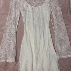 Vit spetsklänning med snörning - Superfin vit kortklänning i spets med långa ärmar och snygg snörning i ryggen. Klänningen har blommigt spetsmönster och är figurnära med lätt utsvängda ärmar. Perfekt för dig som vill sticka ut med en romantisk look. Har dock fått gärna fläckar på sig av en tuschpenna. 