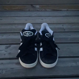Adidas Campus - Svarta Adidas Campus sneakers i mocka med klassiska vita ränder på sidorna och vit logga på plösen. Skorna har snörning, vit kant runt sulan och gummisula i brunt. Perfekta för en streetstyle-look och funkar året runt. Svarta och är i storlek i 35 skriv gärna vid funderingar. Skorna är i väldigt bra skick böjelser bara en lättare rengöring 
