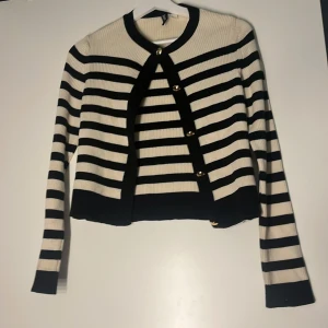 Randig kofta från H&M med guldknappar - Snygg ribbstickad kofta från H&M i svart och vitt med horisontella ränder. Koftan har guldiga knappar framtill och lång ärm. Perfekt för lager-på-lager och ger en klassisk men trendig look. jätte fin mycket bra skick 