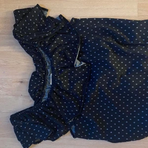 Svart offshoulder blus med småmönster - Snygg svart blus med offshoulder-modell och små vita mönster över hela tyget. Blusen har volangdetaljer upptill och resår vid axlarna för en bekväm passform. Perfekt för dig som gillar trendiga och feminina plagg.