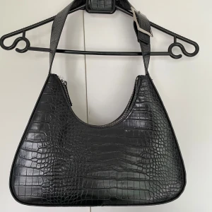 Svart axelväska i crocopräglat skinn - Stilren svart axelväska med crocopräglat mönster och bred axelrem med silverfärgat spänne. Väskan har dragkedja upptill.