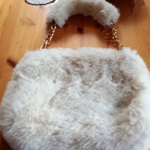 Fluffig beige axelväska med guldkedja - Supermjuk och fluffig axelväska i beige fuskpäls med guldfärgade kedjor som axelrem. Väskan har en chunky look och känns riktigt cozy att bära. Perfekt för dig som vill sticka ut med en trendig accessoar i vinter.