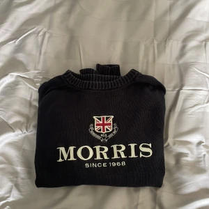 Svart stickad tröja från Morris - Svart stickad tröja från Morris med broderad logga och brittisk flagga framtill. Tröjan har rund halsringning, ribbade muddar och lång ärm. Perfekt för dig som gillar klassisk och stilren design med en preppy touch.