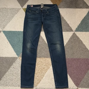 True Religion jeans, mörkblå, stl 28 - Mörkblå jeans från True Religion med ikoniska bakfickor och kontrastsömmar. Klassisk femficksmodell med låg midja och raka ben. Snygga detaljer som logga på bakfickan och flagga vid linningen. Perfekta till sneakers eller boots.