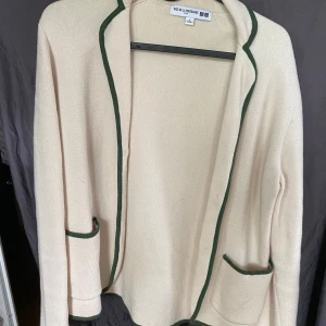Beige cardigan jacka från Uniqlo S - Stilren beige cardigan jacka från Ines de la Fressange x Uniqlo. Jackan har gröna kanter, två fickor framtill och är långärmad. Mjuk stickad känsla som passar perfekt för lager-på-lager. Enkel och snygg att slänga på sig över en t-shirt eller skjorta.