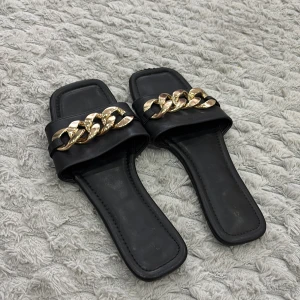 Svarta sandaler med guldkedja, stl 41 - Stilrena svarta sandaler med fyrkantig tå och bred rem framtill, dekorerad med en chunky guldfärgad kedja. Gjorda i skinnliknande material och med platt sula. Perfekta för dig som vill ha en edgy men ändå klassisk look.