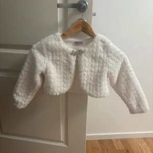Vit pälsjacka från Pretty Baby - Supersöt vit pälsjacka från Pretty Baby i kort modell med lång ärm. Jackan har ett glittrigt spänne framtill och ett fluffigt, mjukt material med struktur. Perfekt för att ge outfiten en extra touch av lyx och cuteness.