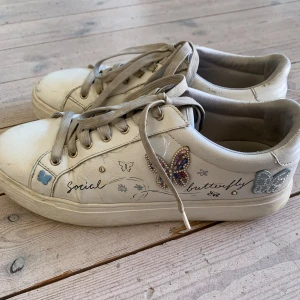Vita sneakers med fjärilsdetaljer - Dessa skor har använts många gånger men är ändå i hyfsat bra skick. Unika vita sneakers med broderade och glittriga fjärilar, texten 'social butterfly' och små pärldetaljer. Skorna har snörning, rund tå och platt sula. Materialet är skinn och de har en cool, personlig vibe med färgglada stenar på fjärilarna.
