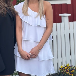 Vit one shoulder volangklänning - Superfin vit kortklänning med one shoulder och flera volanger. Klänningen har en luftig passform och är perfekt för sommarens alla tillfällen. Snyggt axelbandslös design som ger en trendig look. Storlek 152