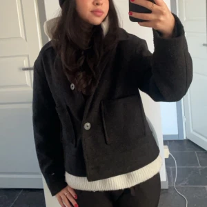 Svart ulljacka med stora fickor - Säljer en svart, kortare jacka i ull med klassisk krage och stora framfickor. Jackan har en rak, loose passform och stängs med knappar framtill. Perfekt för dig som gillar stilren och enkel design med en modern touch.