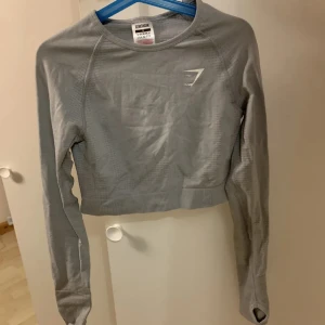 Träningströja - Gymshark - Snygg ljusgrå croppad långärmad träningströja från Gymshark. Tillverkad i stretchigt syntetmaterial som andas, perfekt för träning. Tight stretchig passform och tumhål för extra komfort🩶ANVÄND 1 GÅNG