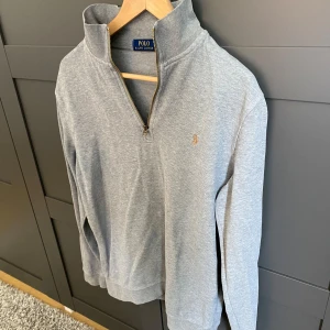 Grå half zip tröja från Polo Ralph Lauren - Snygg grå långärmad tröja från Polo Ralph Lauren med klassisk broderad logga på bröstet. Tröjan har en hög krage och half zip-dragkedja framtill. Tillverkad i mjuk bomull, perfekt för lager-på-lager och chill vardagsstil.