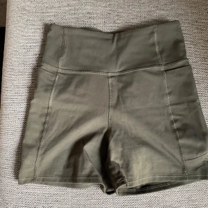 Olivgröna träningstights shorts H&M - Snygga olivgröna träningstights shorts från H&M med medelhög midja och korta ben. De är tillverkade i ett stretchigt material som sitter tight och bekvämt. Perfekta för gymmet eller löprundan, med diskret logga på sidan. Stilrena sömmar och enkel design. De har även en ficka där man kan lägga sin mobil.