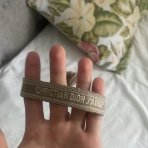 Beige Christian Dior armband - Snyggt beige armband från Christian Dior med broderad logga. Armbandet har en stilren design med diskreta sömmar och är perfekt för att lyfta vilken outfit som helst. Passar dig som gillar exklusiva accessoarer med en clean look.