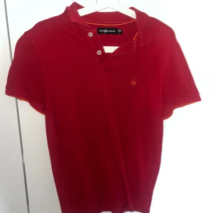 Röd pikétröja från Sail Racing - Röd pikétröja från Sail Racing med klassisk krage och två knappar framtill. Tröjan har korta ärmar och en liten orange logga broderad på bröstet samt en detalj i nacken. Tillverkad i bomull, perfekt för en avslappnad och sportig stil.