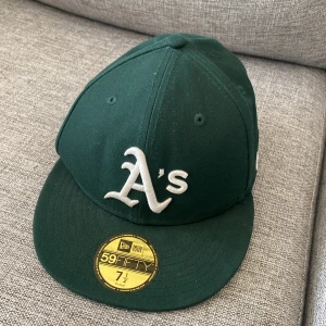 Grön Oakland A's keps New Era 59FIFTY - Säljer en mörkgrön Oakland Athletics keps från New Era, modell 59FIFTY. Kepsen har broderad A's-logga framtill, 1989 World Series-märke på sidan och klassisk rak skärm. Tillverkad i bomull och polyester, perfekt för dig som gillar sportig streetstyle.