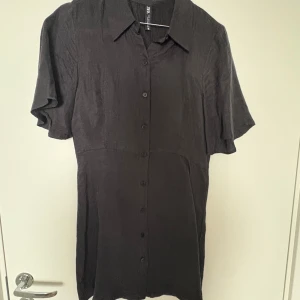 Svart skjortklänning från Zara - Svart skjortklänning från Zara i storlek S. Klänningen har korta ärmar, klassisk krage och knappar hela vägen framtill. Tillverkad i ett lätt, vävt material som ger en stilren och enkel look. Perfekt för dig som gillar minimalistisk stil.