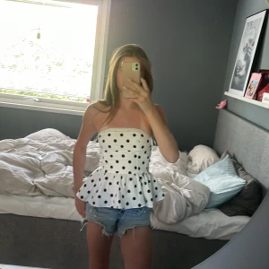 Vit prickig bandeau tubtopp - Supersöt vit bandeau tubtopp med svarta prickar och volang nertill. Toppen är axelbandslös och har en figurnära passform upptill som blir lite utsvängd vid midjan. Perfekt för varma dagar och ger en riktigt fräsch sommarkänsla.