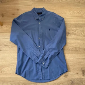 Blå skjorta från Ralph Lauren - Säljer en stilren blå skjorta från Ralph Lauren i slim fit-modell. Skjortan har klassisk krage, knappar framtill och broderad logga på bröstet. Tillverkad i mjuk bomull och har långa ärmar. Perfekt för dig som gillar en clean och tidlös look.