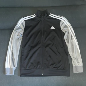 Svart Adidas track jacket med grå ärmar - Snygg svart Adidas track jacket med grå ärmar och klassiska vita ränder längs axlarna. Jackan har dragkedja hela vägen, ståkrage och två fickor framtill. Adidas-logga på bröstet. Perfekt för en sportig look.