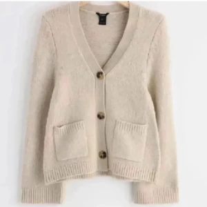 Beige stickad kofta med knappar - Mysig beige stickad kofta med stora knappar framtill och två fickor. Koftan har v-ringning, ribbade muddar och en relaxed passform som passar perfekt till jeans. Enkel och stilren design som är lätt att matcha.