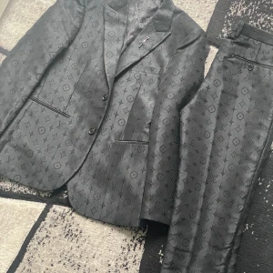 Svart Louis Vuitton kostym med monogram - Exklusiv svart kostym från Louis Vuitton med ikoniskt monogrammönster över hela tyget. Kavajen har klassisk passform, två knappar och snygga fickdetaljer. Byxorna är raka och matchar perfekt. Materialet har en lyxig glans och känns riktigt premium.