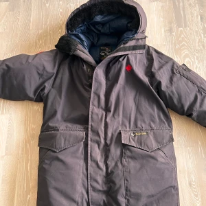 Svart dunjacka från Canada Goose - Svart vintage dunjacka från Canada Goose med huva och stora fickor framtill. Jackan har en röd lönnlöv-detalj på bröstet, reflexband på ryggen och broderad logga. Fodrad insida i blått och svart, perfekt för kalla dagar. Jackan är från 1990-talet men är som ny.