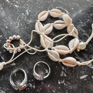 Zend Details snäckhalsband & coola ringar - Säljer smycken från Zend Details. Ett vitt snäckhalsband med pärldetalj, en silverfärgad ring med blomma i vitt och gult, en glittrig ring med smiley och en öronring med pärlor och gulddetaljer. 