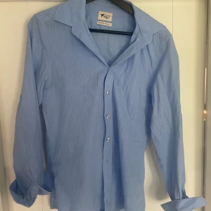 Ljusblå skjorta från AC&Co, slim fit - Snygg ljusblå skjorta från AC&Co i slim fit-modell. Skjortan har klassisk krage, långa ärmar och knäppning framtill. Tillverkad i ett lätt och svalt bomullstyg som passar perfekt till både jeans och kostymbyxor.