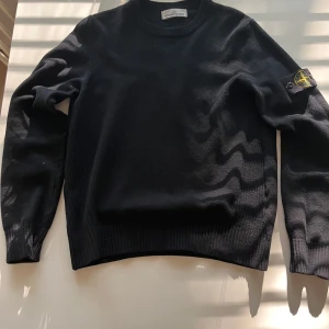 Svart ulltröja från Stone Island - Svart långärmad tröja från Stone Island i mjuk ull med klassisk rund hals och ribbade muddar. Ikonisk logotyp-patch på vänster ärm som fästs med knappar. Perfekt för dig som gillar stilren och clean streetwear.