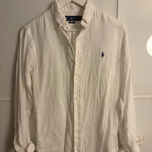 Vit linneskjorta Ralph Lauren - Vit långärmad skjorta från Ralph Lauren i slim fit-modell. Tillverkad i linne med klassisk krage, knäppning framtill och ikonisk blå broderad logga på bröstet. Perfekt för en clean och stilren look.
