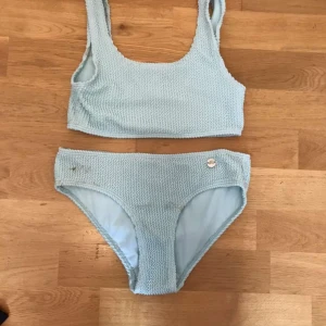 Ljusblå bikini från Kappahl - Supersöt ljusblå bikini från Kappahl i sportig modell med breda axelband och hög midja på underdelen. Bikinin har en ribbad struktur och en liten metallknapp på trosan. Perfekt för strandhäng och poolparty!