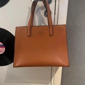 Brun handväska från Kstyle - Snygg brun handväska från kstyle slätt skinn med stilren, fyrkantig form. Väskan har två rejäla handtag, dragkedja baktill och flera fack inuti för smart organisering. Perfekt för dig som vill ha en klassisk och tidlös look.