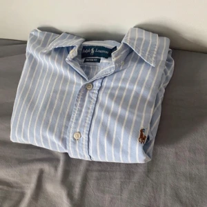 Blåvit randig skjorta från Ralph Lauren - Snygg blå skjorta med vita ränder från Ralph Lauren. Klassisk krage, långa ärmar och knäppning framtill. Ikoniska broderade polospelaren på bröstet. Skjortan är i bomull och har en normal passform, storlek S