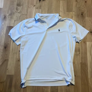 Vit pikétröja från Ralph Lauren - Klassisk vit pikétröja från Ralph Lauren med broderad marinblå logga på bröstet. Tröjan har korta ärmar, krage och två knappar framtill. Tillverkad i mjukt och svalt material som passar perfekt till sommaren.