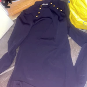 Mörkblå klänning med guldknappar - Snygg mörkblå klänning med långa ärmar och dekorativa guldknappar vid axlarna. Stilren och enkel design som ger en cool och elegant vibe. Perfekt för dig som gillar detaljer och vill sticka ut lite extra.