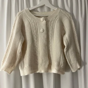 Vit stickad kofta från 157 - Supermysig vit stickad kofta från 157 med flätstickade detaljer och två stora knappar framtill. Koftan har rund halsringning, ribbade muddar och en loose fit som ger en chill vibe. Ärmarna är tre kvarts. Perfekt att slänga på sig när det är kallt ute!