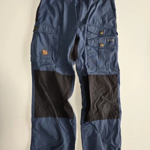 Fjällräven blå cargopants barn - Blå cargopants från Fjällräven med svarta förstärkningar på knän och bak. Byxorna har flera fickor med tryckknappar och klassisk G-1000 patch. Perfekta för äventyr och outdoor, tillverkade i slitstarkt bomullsmaterial.