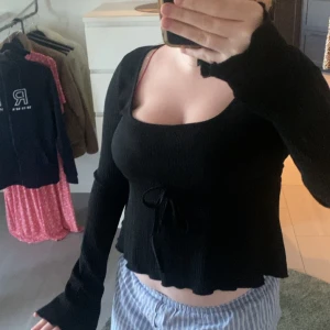 Svart croppad topp från Nelly - Svart långärmad croppad topp från Nelly med ribbad struktur och knytning framtill. Toppen har en fyrkantig urringning och långa ärmar med lätt utsvängda ärmslut. Perfekt för dig som gillar en enkel men ändå trendig look. Och använt 1 gång. 